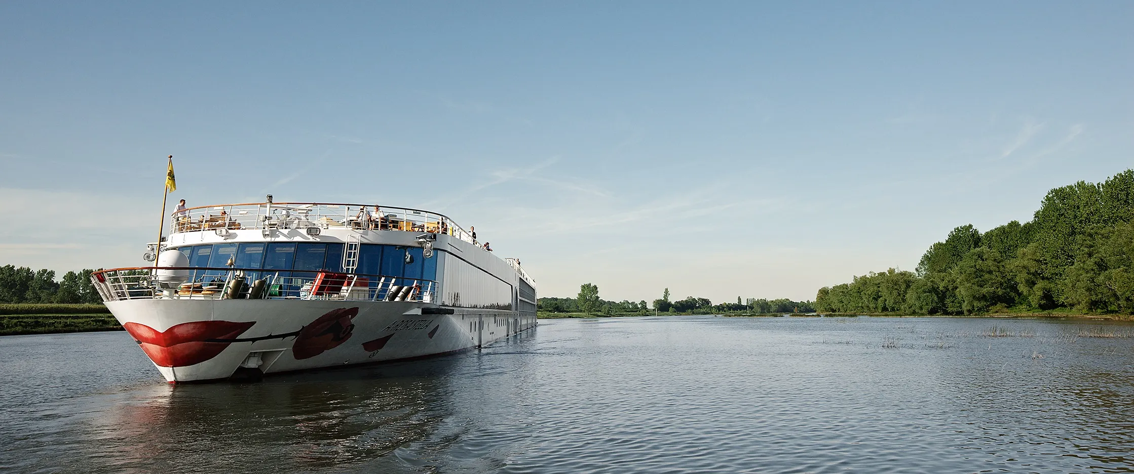 Flusskreuzfahrtschiff auf ruhigem Fluss, sonniger Tag, Naturkulisse.