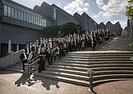 Orchester auf Treppe vor modernem Gebäude, sonnige Stimmung.