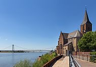 Kirche am Flussufer mit Brücke, sonniger, klarer Himmel.