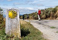 Wegweiser mit gelber Muschel, Wanderer auf Jakobsweg, sonnig.