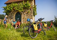 Radreise Pfälzer Weinsternfahrt, Radpause vor historischem Gebäude bei der Pfälzer Weinsternfahrt., Radfahrer entspannen vor einer malerischen, bewachsenen Steinhütte.