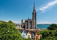 Cork (Route 2), St. Colman's Cathedral in Cobh mit bunten Häusern im Vordergrund., St. Colman's Cathedral in Cobh, bunte Häuser und blauer Himmel.
