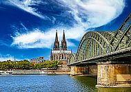 Kölner Dom und Hohenzollernbrücke am Rhein unter blauem Himmel.