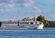 MS Olympia am Fahren, MS Olympia auf ruhiger Flussfahrt unter blauem Himmel., MS Olympia auf ruhiger Flussfahrt vor malerischer Kulisse.