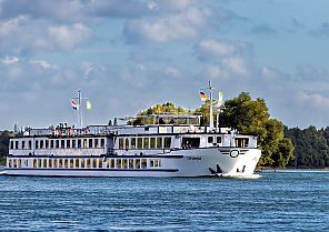 MS Olympia am Fahren, MS Olympia auf ruhiger Flussfahrt unter blauem Himmel., MS Olympia auf ruhiger Flussfahrt vor malerischer Kulisse.